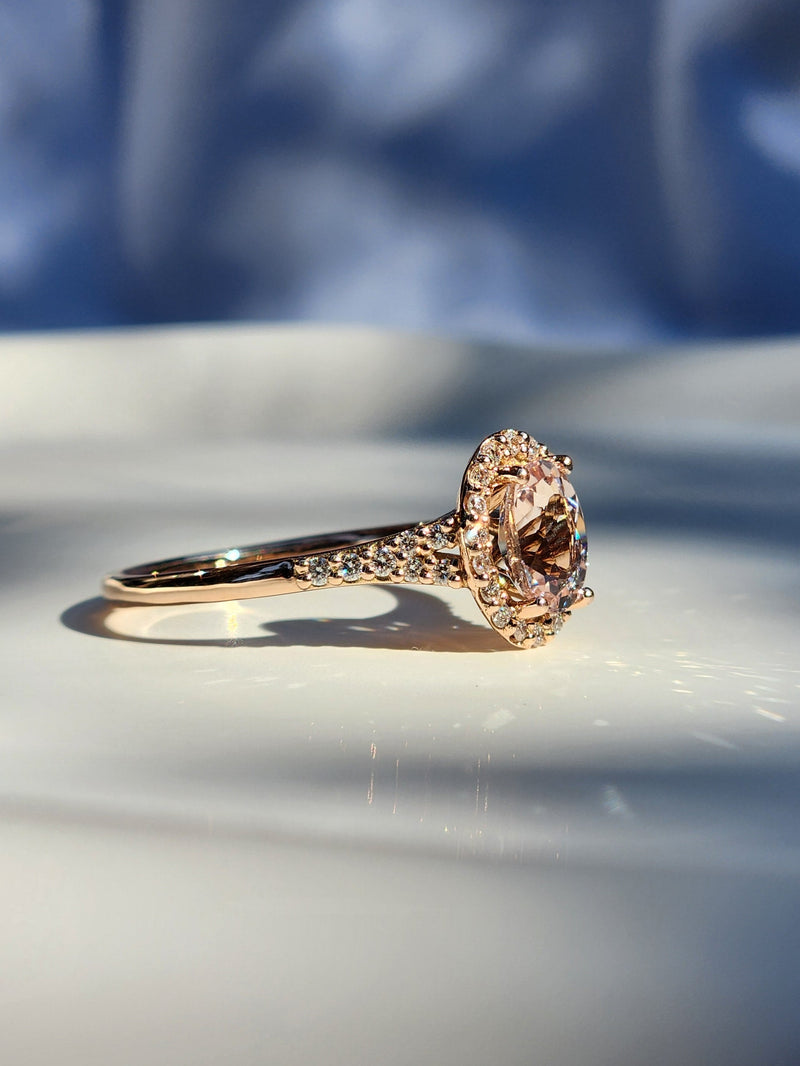 A rose gold engagement ring with a large oval-cut pink gemstone and small accent stones, resting on a white surface with soft blue shadows and a blurred blue background.
