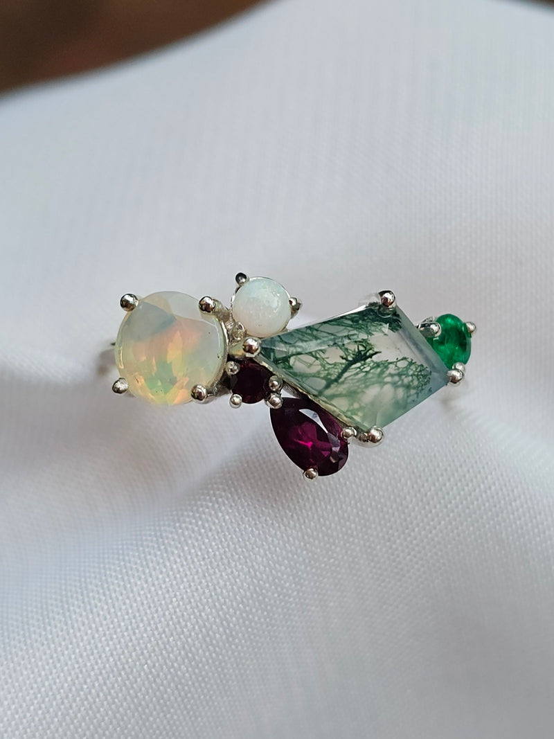 A cluster ring featuring a large iridescent opal, a moss agate, a dark purple garnet marquise gemstone, and a small emerald round gemstone, set in white gold metal on a white fabric background.