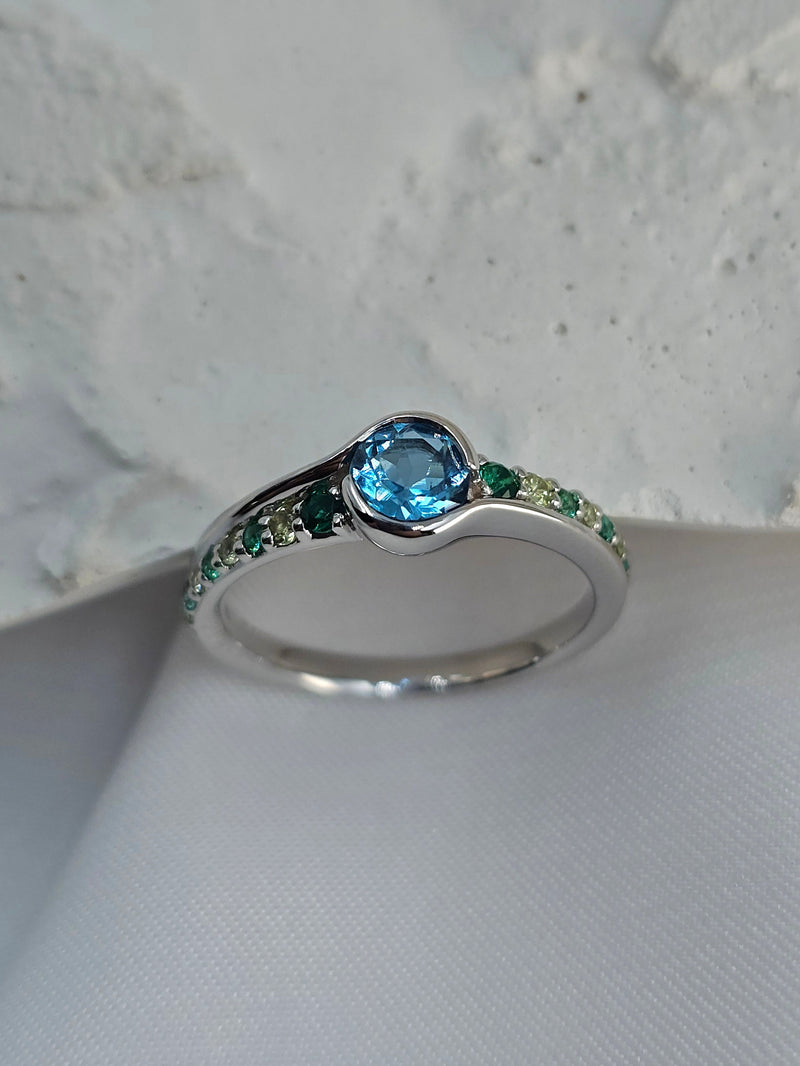 A white gold ring featuring a round-cut light blue topaz center stone and a channel-set row of small green gemstones along the band, displayed on a textured white surface.